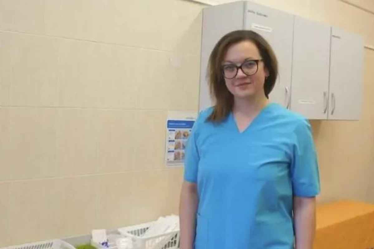Moteris su trumpomis rudos spalvos plaukais ir akiniais vilki mėlynos spalvos medicininę uniformą gydymo įstaigos patalpoje.